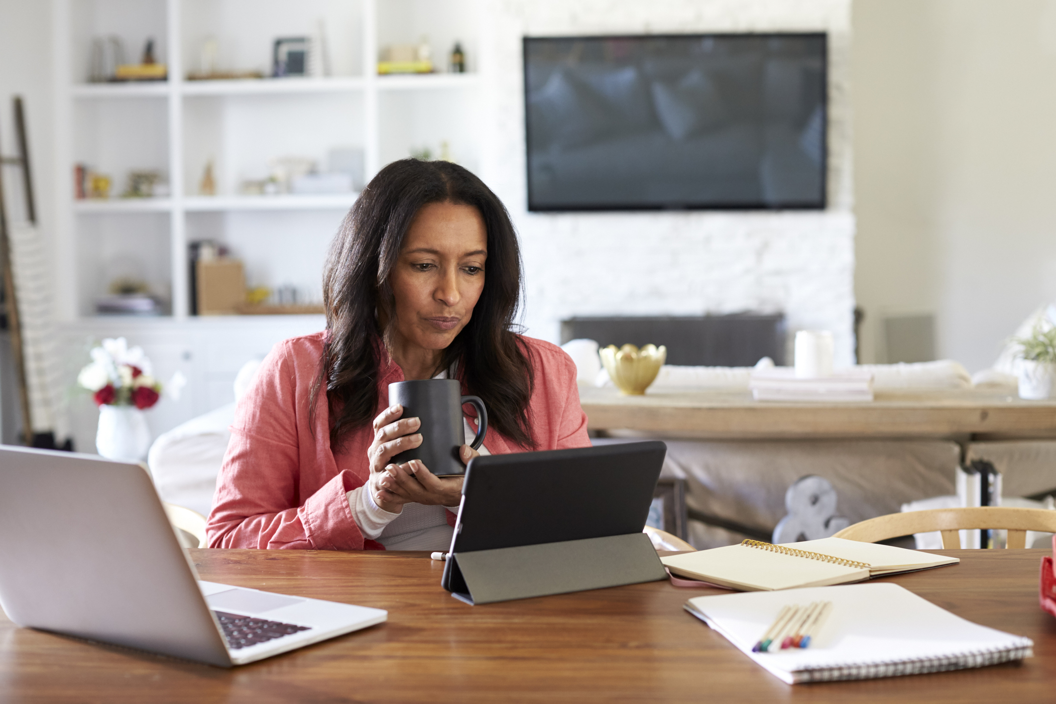 Female working from home on a laptop and tablet device whilst drinking a hot drink. She is sat at a table with her living room in the background
