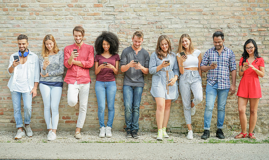 Nine people stood in a line, leaning against a brick wall and using their mobile phones