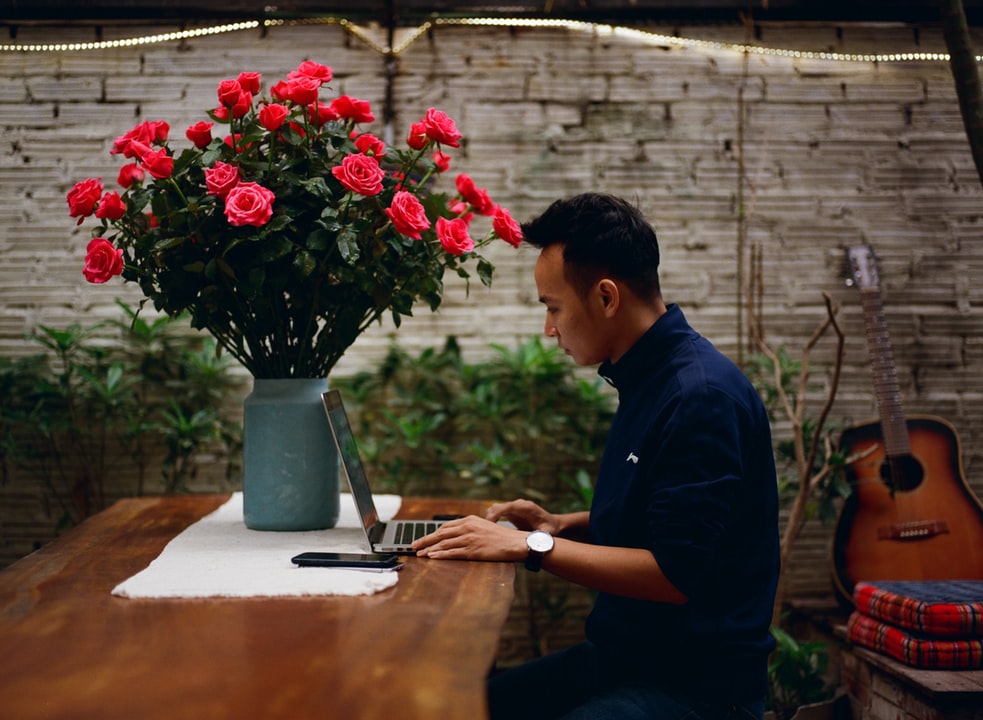 Male sitting at a desk next to a large vase of pink flowers. He is wearing a dark blue shirt and using a laptop