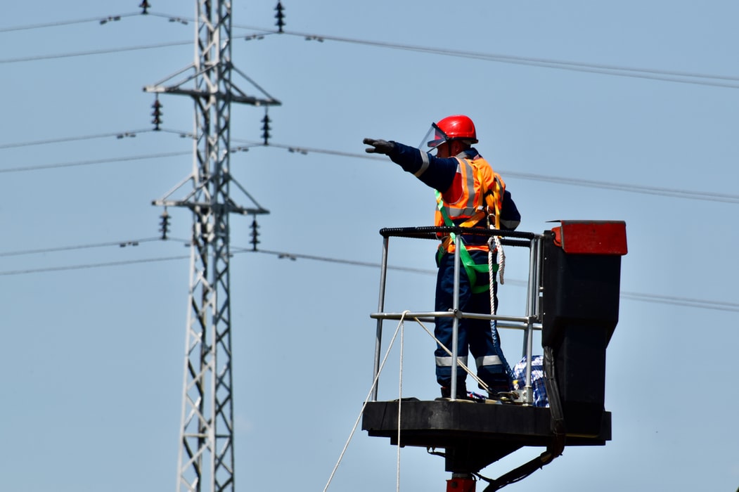 An electrician standing on an extended cherry picker raising his arm towards a telegraph pole in the distance