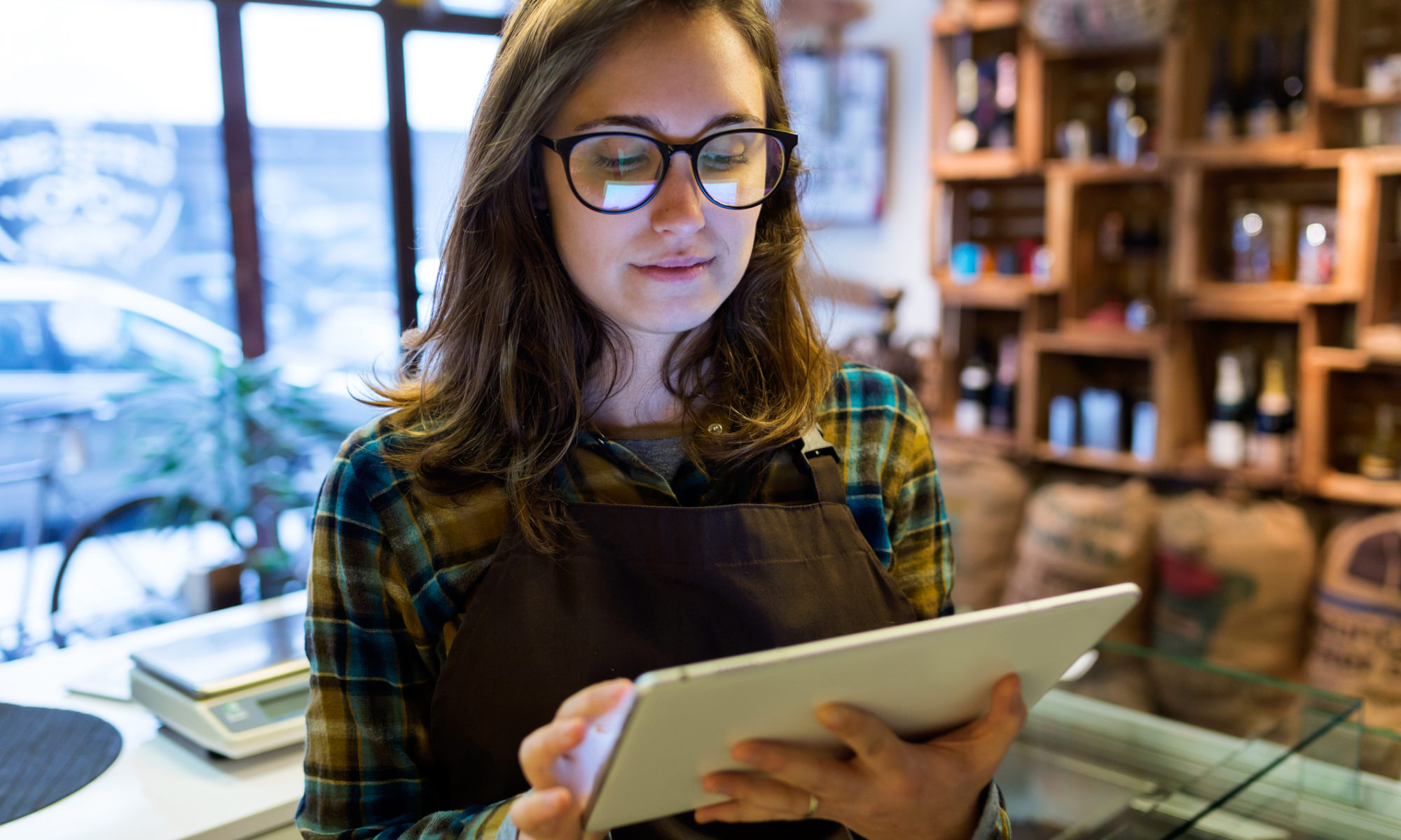 Female small business owner stood in the middle of her shop updating her online store via a tablet device. She is wearing dark green clothing and glasses