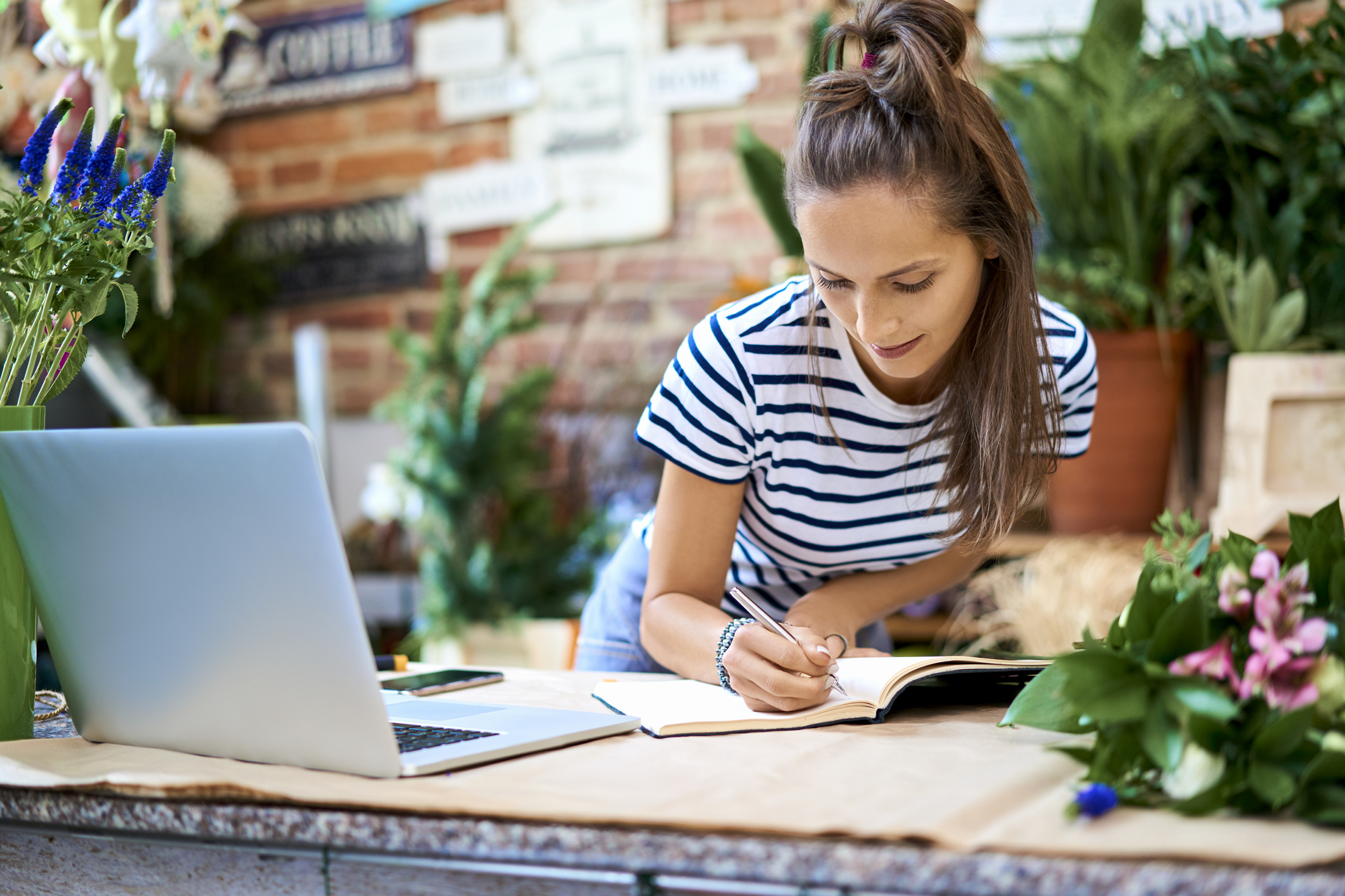 Young, female florist writing out the costs of launching her new brand in her flower shop alongside her laptop. She is wearing a black and white striped top.