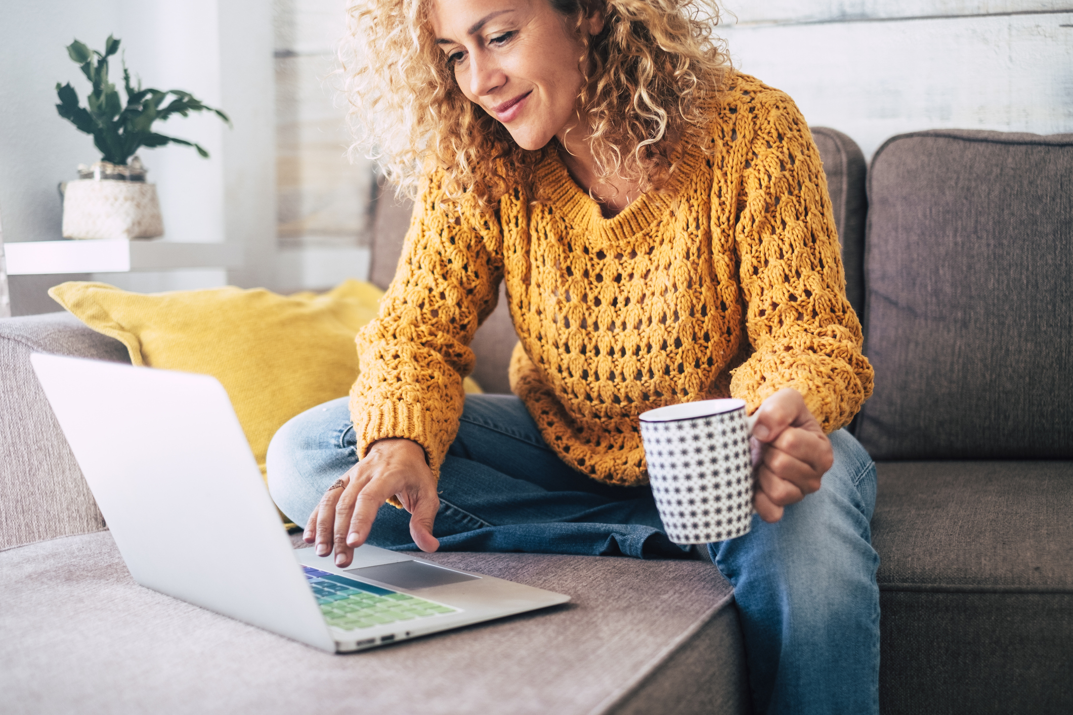 Female small business owner sat on her sofa at home with a hot coffee planning her SEO strategy on her laptop. She is casually dressed and smiling