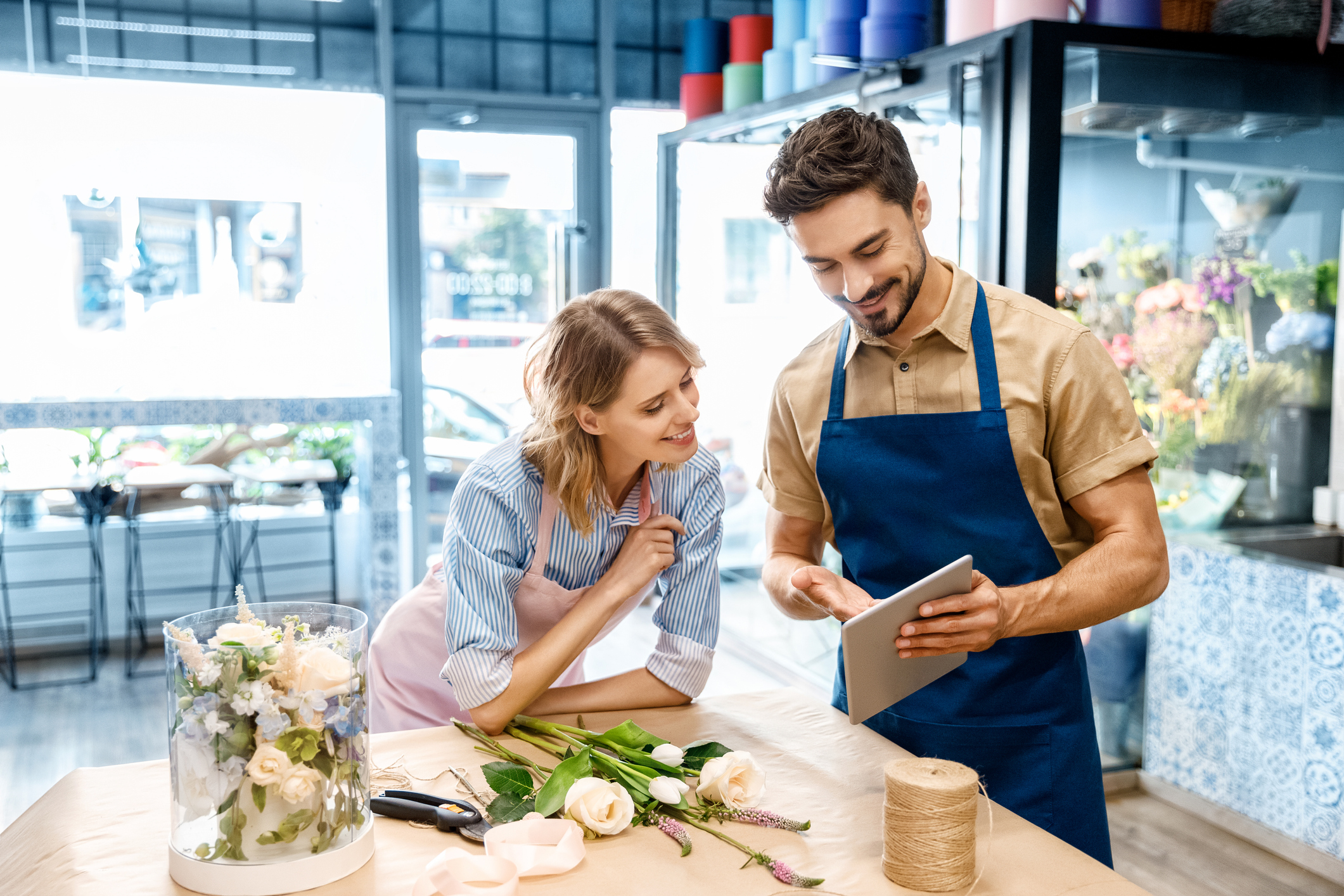 A male and a female business owners stood in a shop looking at a tablet device. Both are wearing aprons and smiling