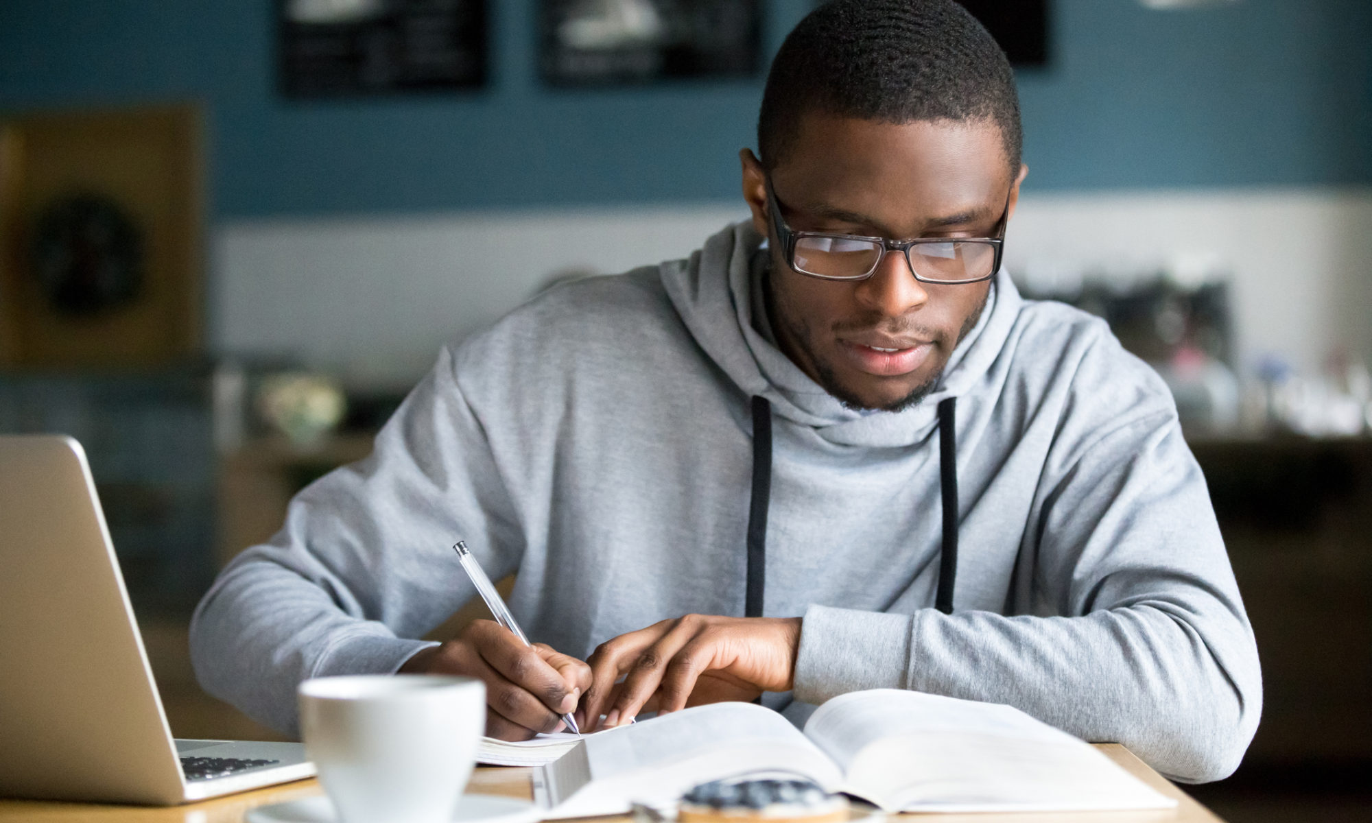 Young black male entrepreneur studying marketing strategies for his new online store. He is sat at a desk with a coffee and laptop, writing in a notebook