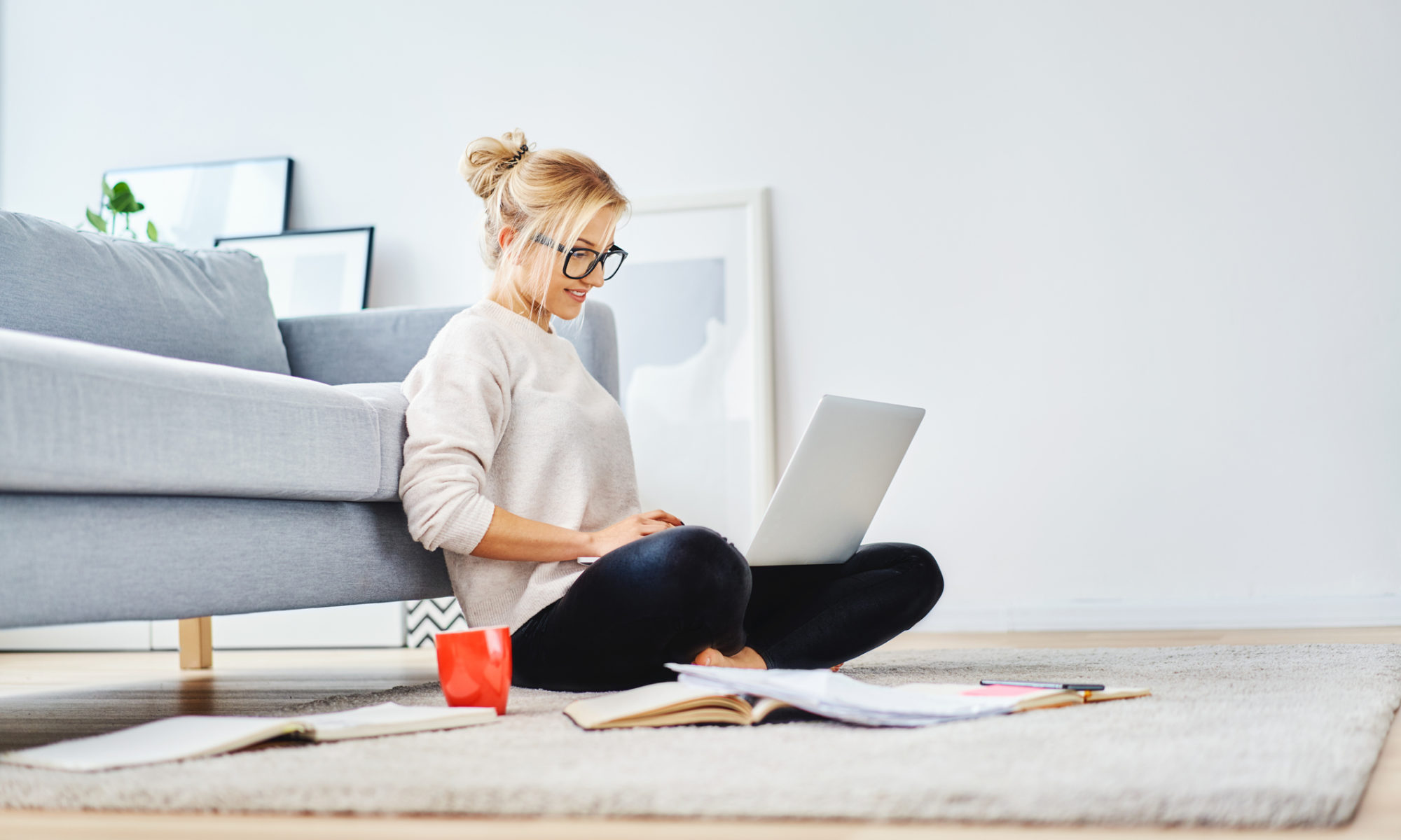 Female small business owner working at home and using her laptop to help her business rank higher on Google. She is sat on the floor of her living room, leaning against a grey sofa.