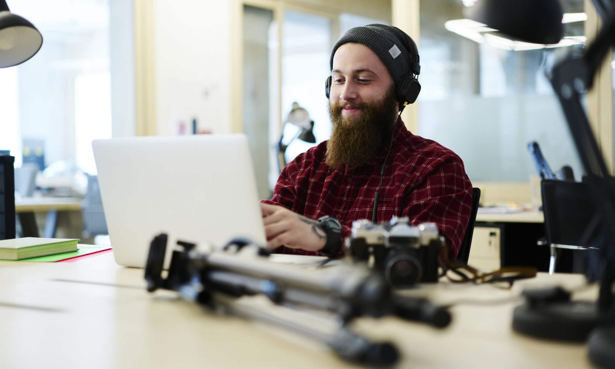 Caucasian male with a long beard is sat at a desk in front of a laptop editing photos with camera equipment surrounding the laptop
