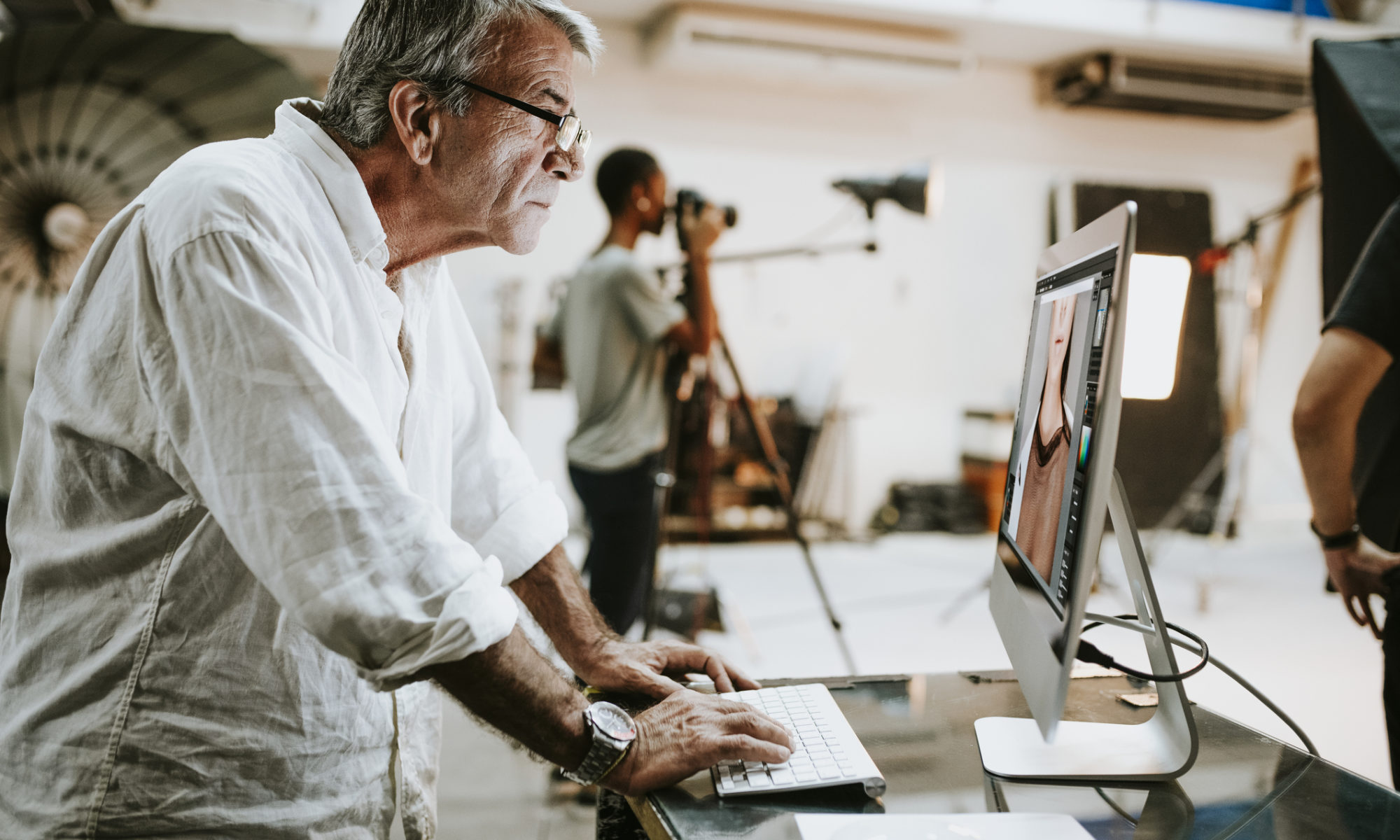 A male photographer wearing a loose, casual white shirt is stood at a desktop computer editing images whilst a photoshoot is going on in the background