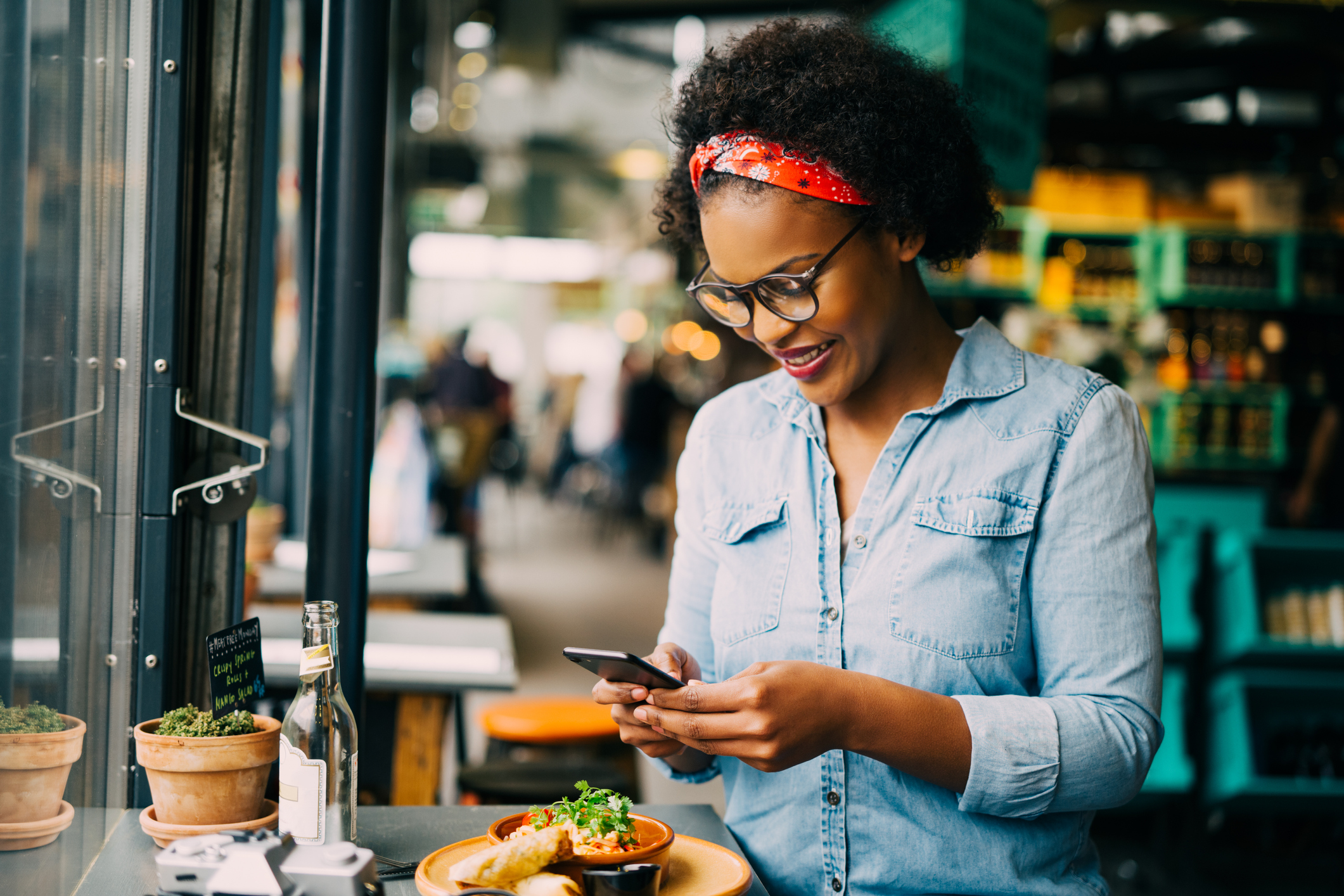Black female food blogger taking a photo in a cafe wearing a light blue shirt and red hair bandana