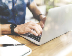 Male wearing a short sleeved blue shirt is sat at a desk typing on this laptop. He has a black watch on his right wrist and a notebook and pen is next to the laptop