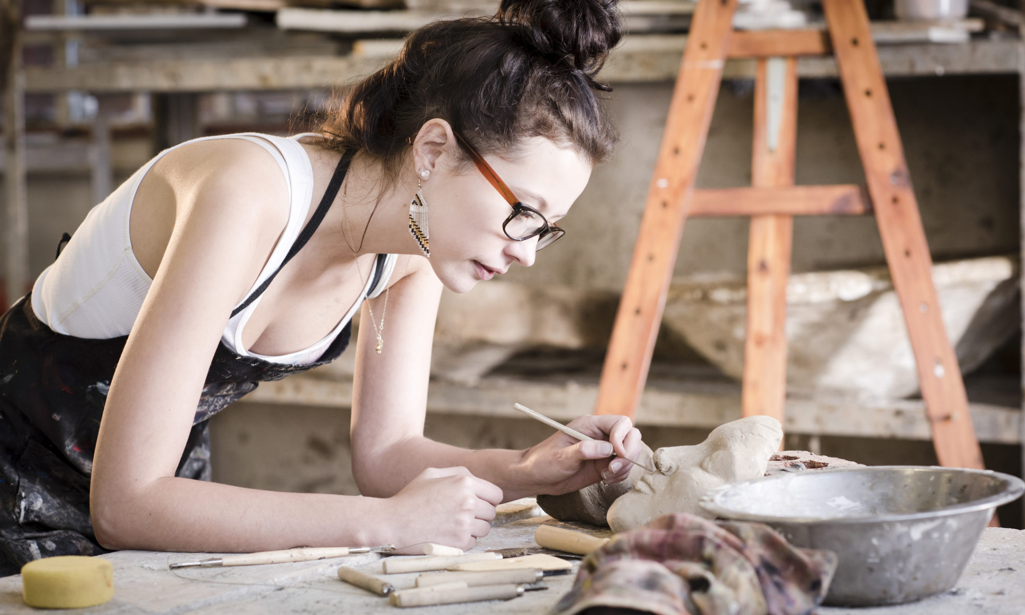 A caucasian female potter leaning over a workbench adding finishing touches to a piece of pottery. She is wearing a white strap top with a black apron on top