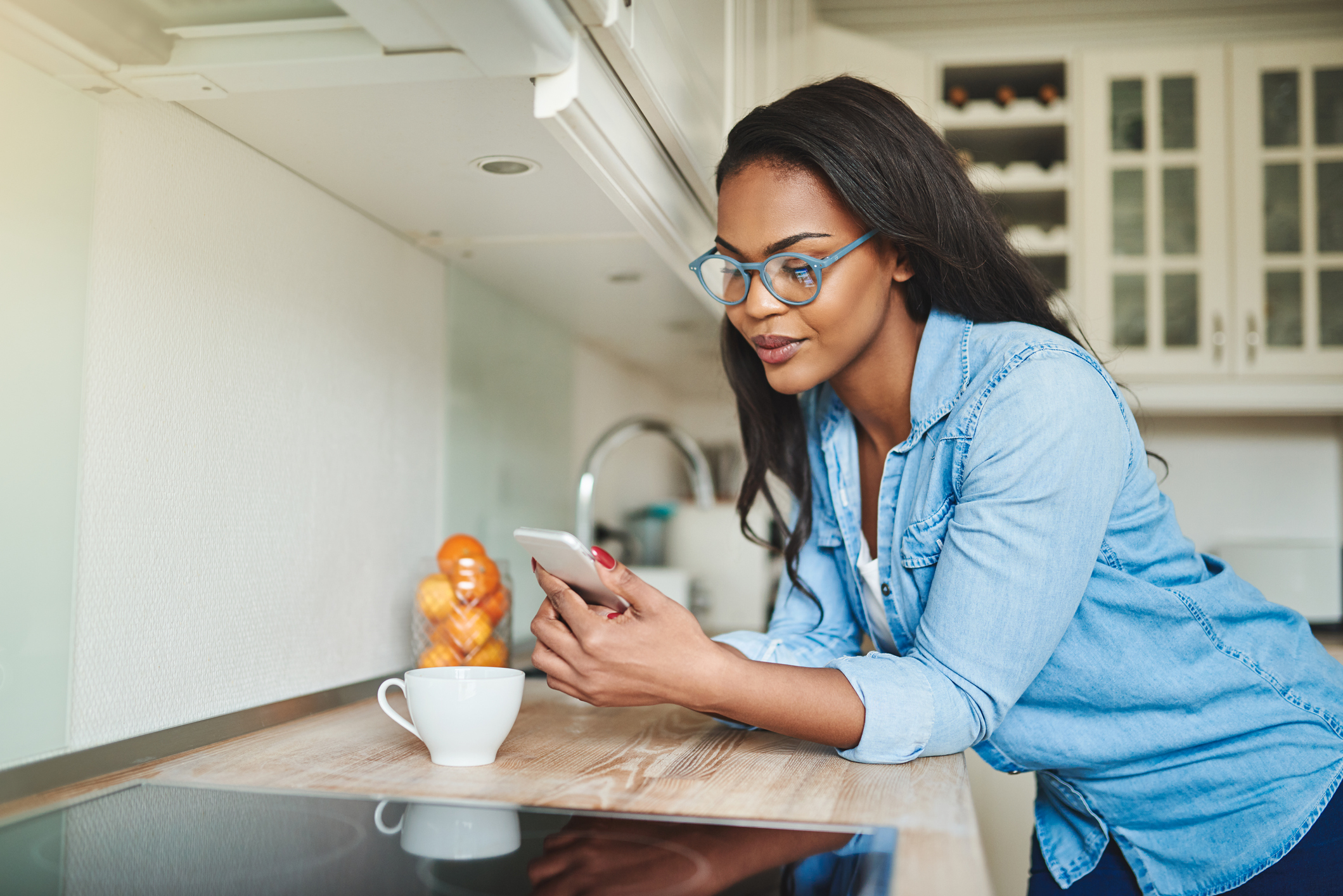 Dual heritage female business owner using her phone to update her website's SEO. She is casually dressed standing in her kitchen wearing glasses and drinking coffee