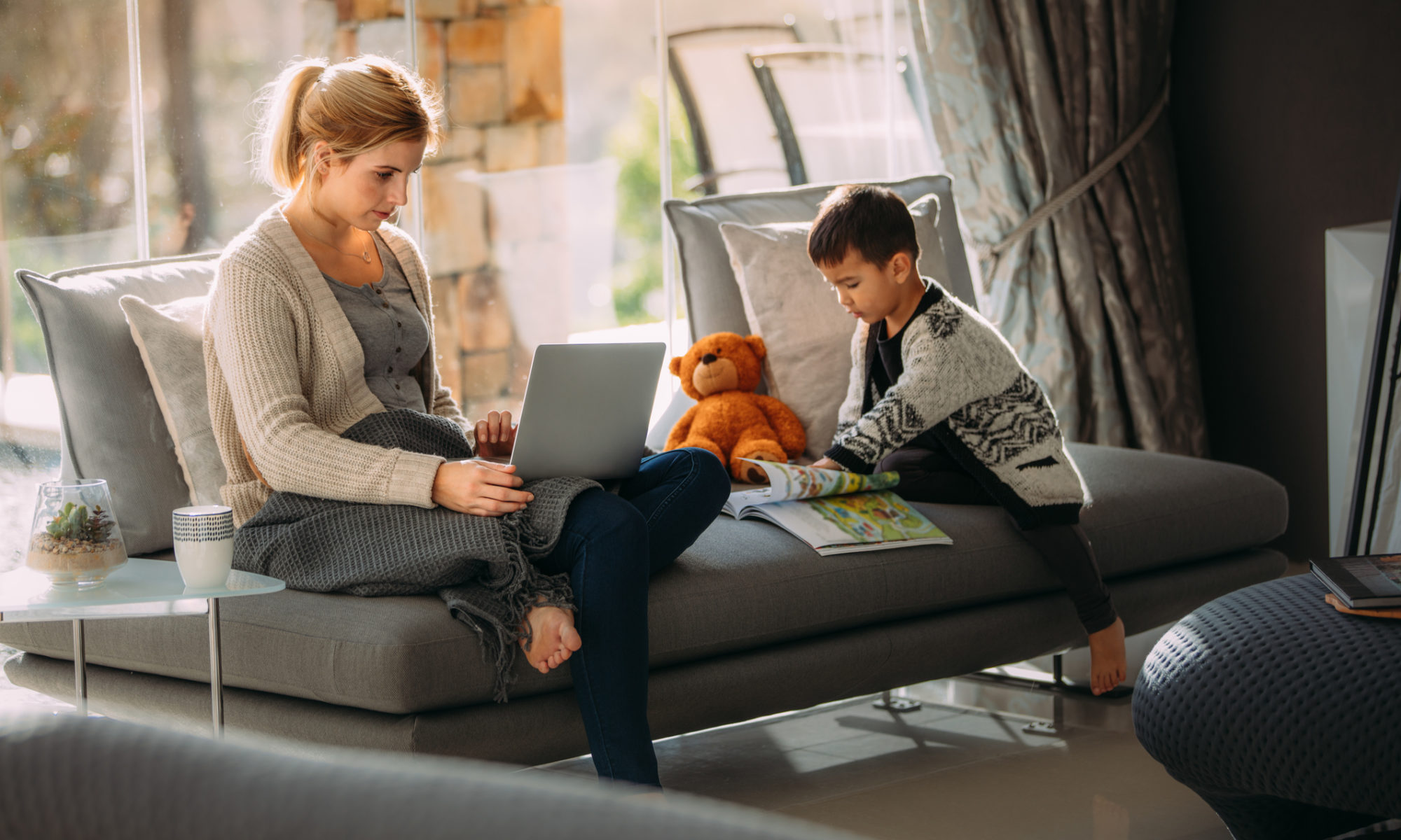 Working woman on laptop trying to make her website look more professional with her son reading a storybook. Both sitting on a grey sofa in a living room.