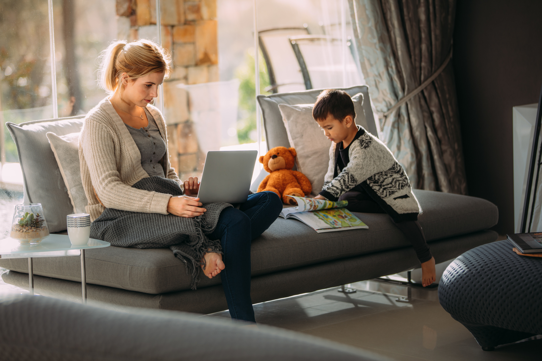 Working woman on laptop trying to make her website look more professional with her son reading a storybook. Both sitting on a grey sofa in a living room.