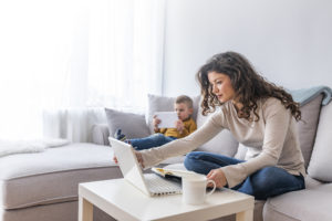 Mother working on laptop at home, while her child using tablet. Boy and mother sat on the sofa. The Mother is managing her business on her laptop during the Covid-19 crisis