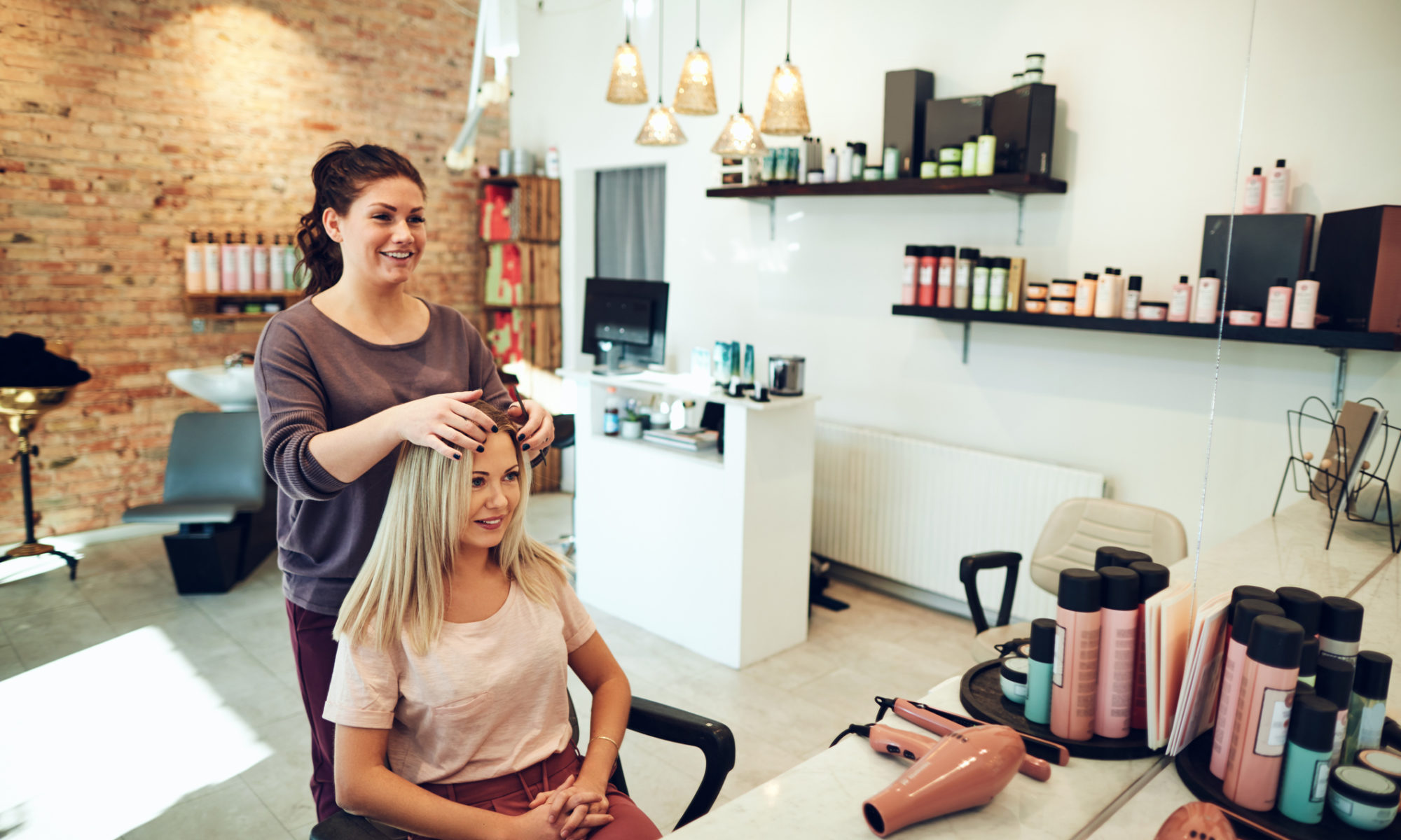 Young blonde woman smiling and looking at her reflection in a mirror while sitting in a salon chair discussing the costs of a new salon website