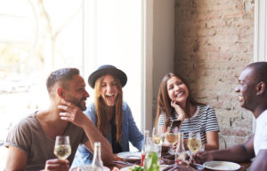 Diverse small group of four attractive laughing friends having wine and a meal together in restaurant talking about how much it cost to make a website for a small restaurant