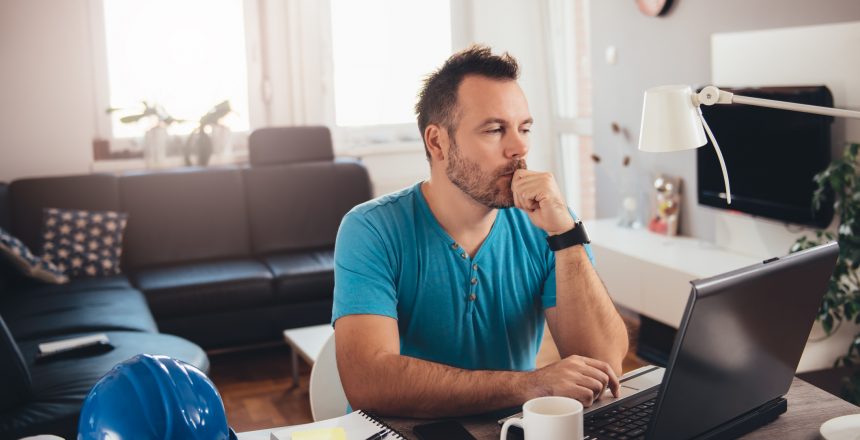 Male business owner working from home and using his laptop to update this Go Sitebuilder website. He is wearing a blue v-neck t-shirt, sitting at a desk that has a mug, notebook and hard hat on.