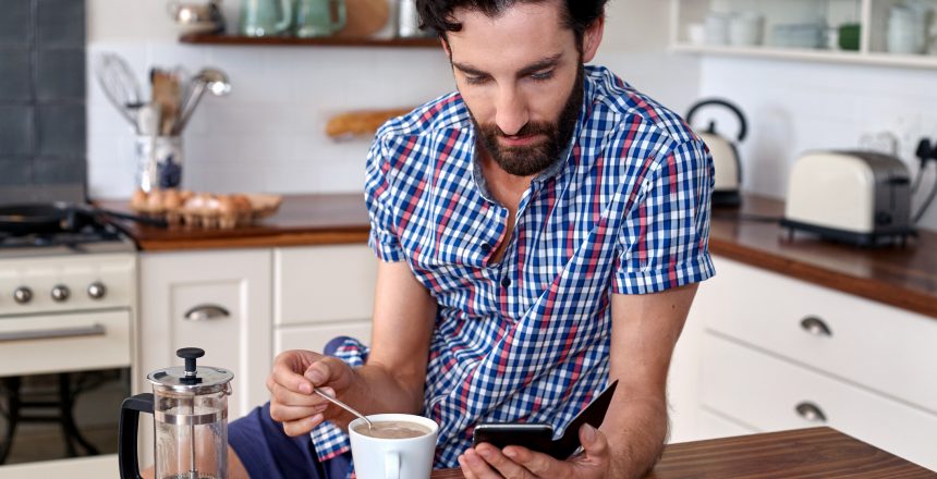 Male wearing a blue, white and red checkered shirt is standing in his kitchen looking at his phone and making a coffee