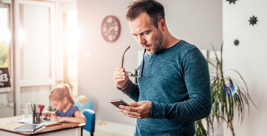 Male looking at mobile phone whilst holding reading glasses. He is working from home and his young daughter can be seen eating at a table in the background.