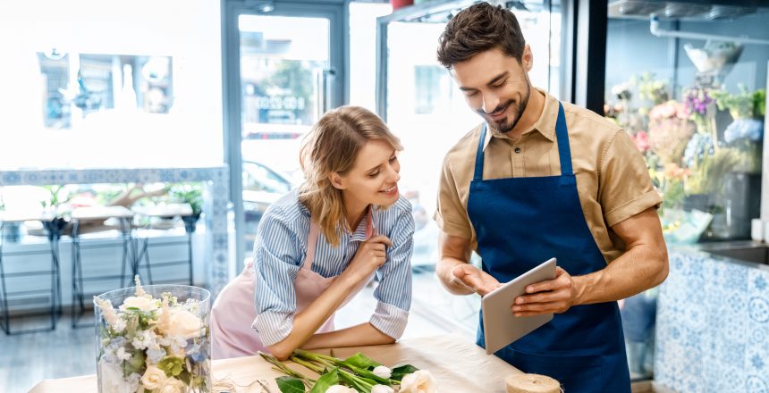 A male and a female business owners stood in a shop looking at a tablet device. Both are wearing aprons and smiling