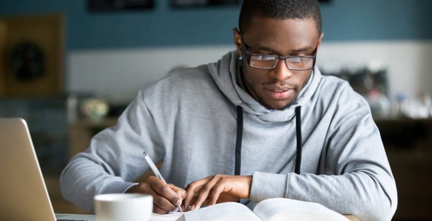 Young black male entrepreneur studying marketing strategies for his new online store. He is sat at a desk with a coffee and laptop, writing in a notebook