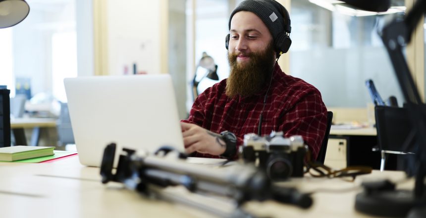 Caucasian male with a long beard is sat at a desk in front of a laptop editing photos with camera equipment surrounding the laptop