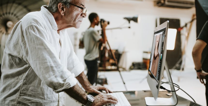A male photographer wearing a loose, casual white shirt is stood at a desktop computer editing images whilst a photoshoot is going on in the background