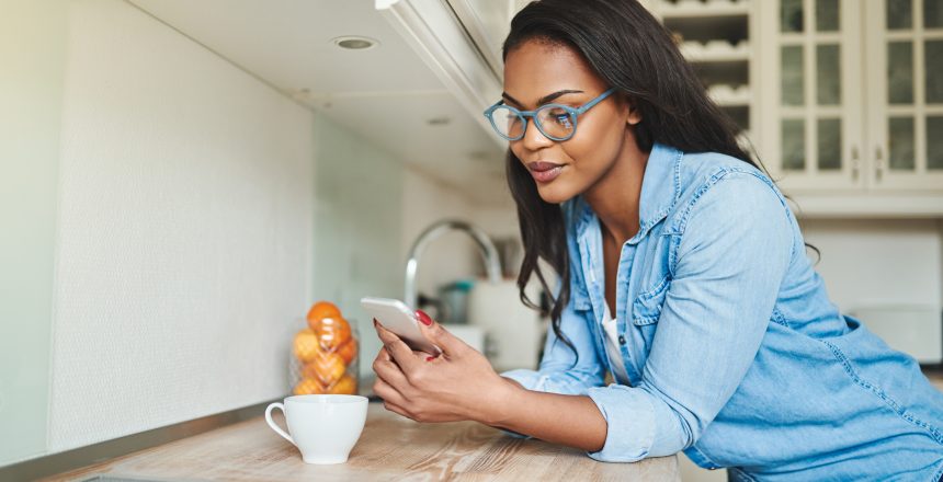 Dual heritage female business owner using her phone to update her website's SEO. She is casually dressed standing in her kitchen wearing glasses and drinking coffee