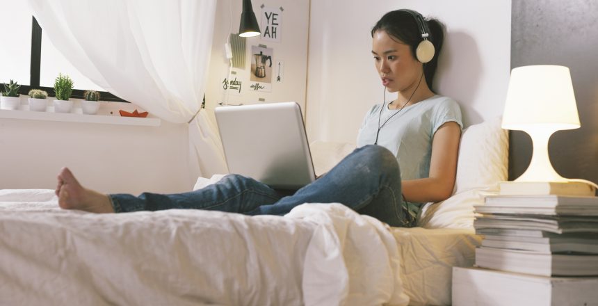 Young female sitting on a mattress in a trendy bedroom on her laptop and listening to music through headphones