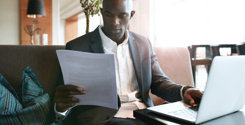 Black male lawyer dressed in a suit is sitting on his sofa at home working on his laptop and viewing some paperwork that he's holding in his hand