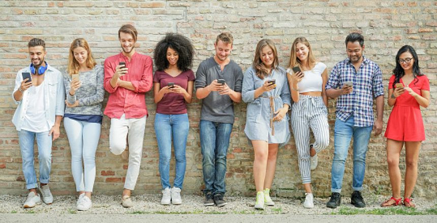 Nine people stood in a line, leaning against a brick wall and using their mobile phones