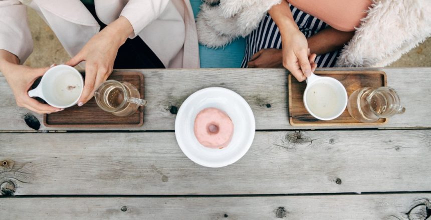 Birds eye view of two females sat down at a table drinking tea. A doughnut is also on the table.