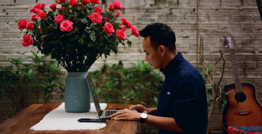 Male sitting at a desk next to a large vase of pink flowers. He is wearing a dark blue shirt and using a laptop