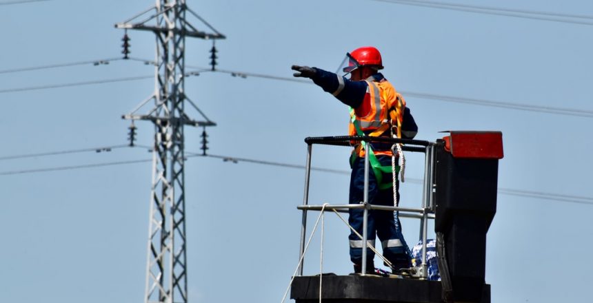 An electrician standing on an extended cherry picker raising his arm towards a telegraph pole in the distance